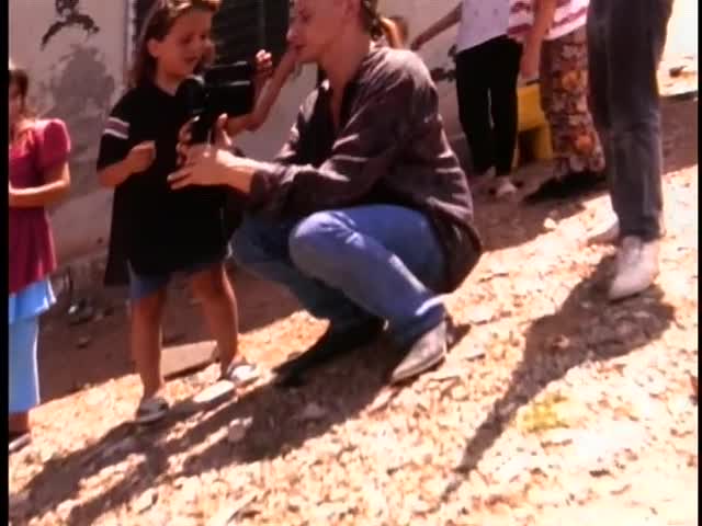 A man crouches down, holding a camera as a young girl speaks into it. Other children and adults stand nearby, watching the scene unfold.

