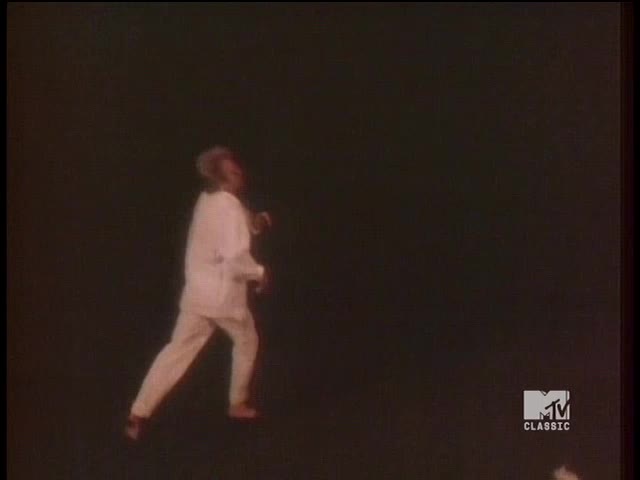 A man in a white suit strides across the dark space, his movements fluid. The MTV Classic logo sits in the corner, a reminder of the music video playing on the screen.
