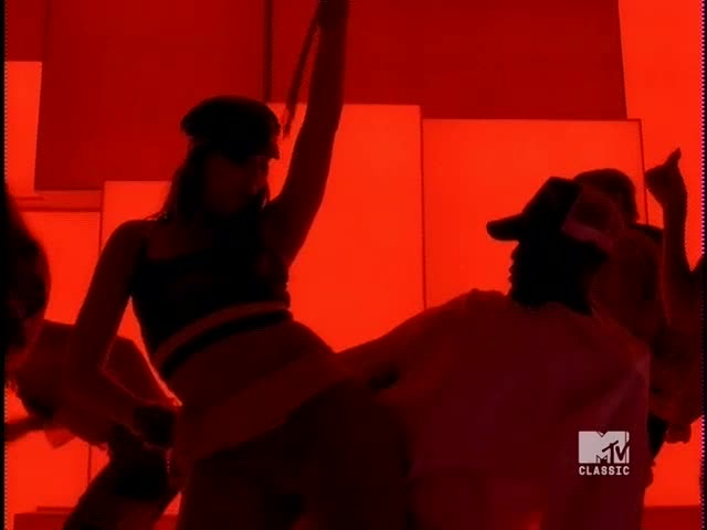 The dancers are silhouetted against a red background, moving in a dimly lit space. A woman in a cap raises her arm, while a man in a hat moves in front of her.
