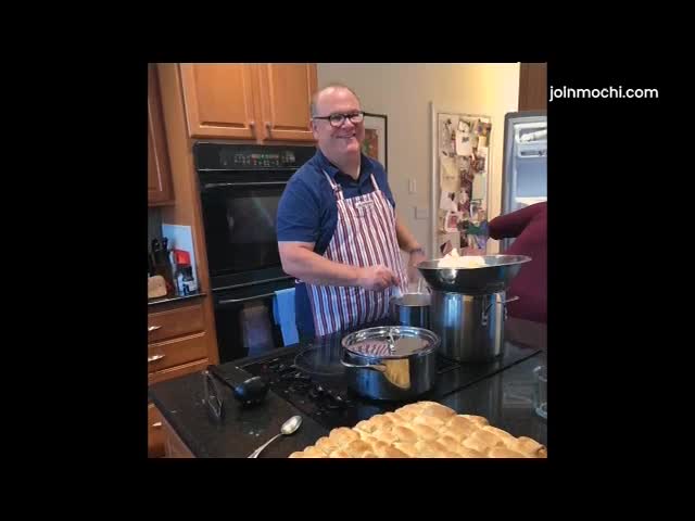A man in an apron stirs something in a pot on the stove. A tray of golden-brown rolls sits in the foreground.
