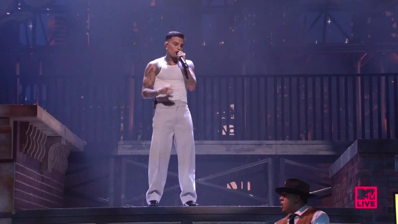 On a stage designed to look like a city street, a man in a white tank top and slacks sings into a microphone. Below, another performer in a fedora and vest seems to be singing backup.
