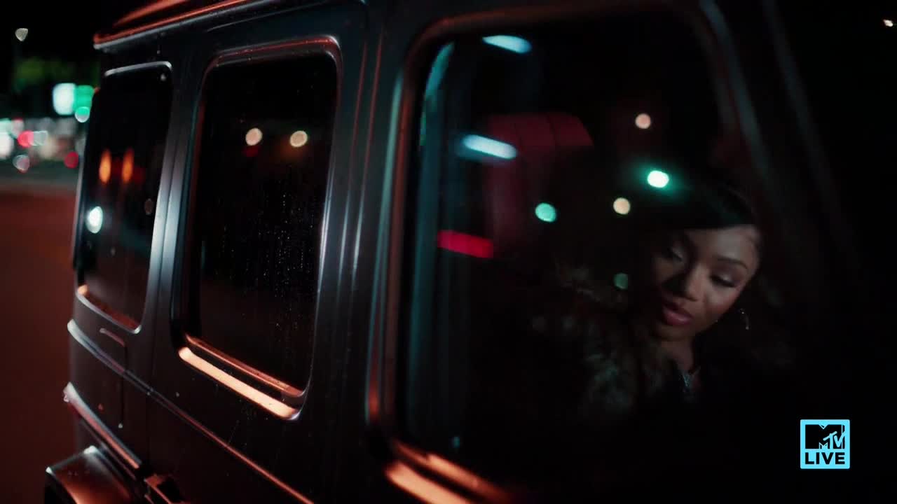 Streetlights streak past the windows of a black SUV as it moves. Inside, a woman looks down, illuminated by the glow of the city.
