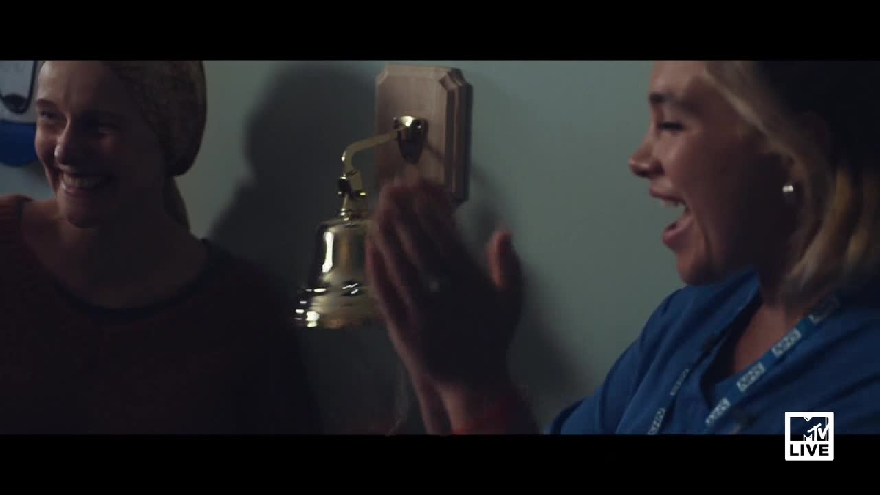 A woman in a blue shirt is clapping excitedly, her mouth wide open. Beside her, another woman smiles as they stand near a shiny, golden bell mounted on the wall.
