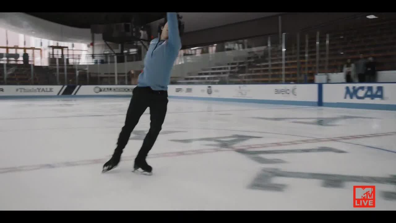 A skater in a light blue shirt and black pants is mid-air, arms raised above his head. The ice beneath him is marked with large letters, and the MTV Live logo is visible in the corner.
