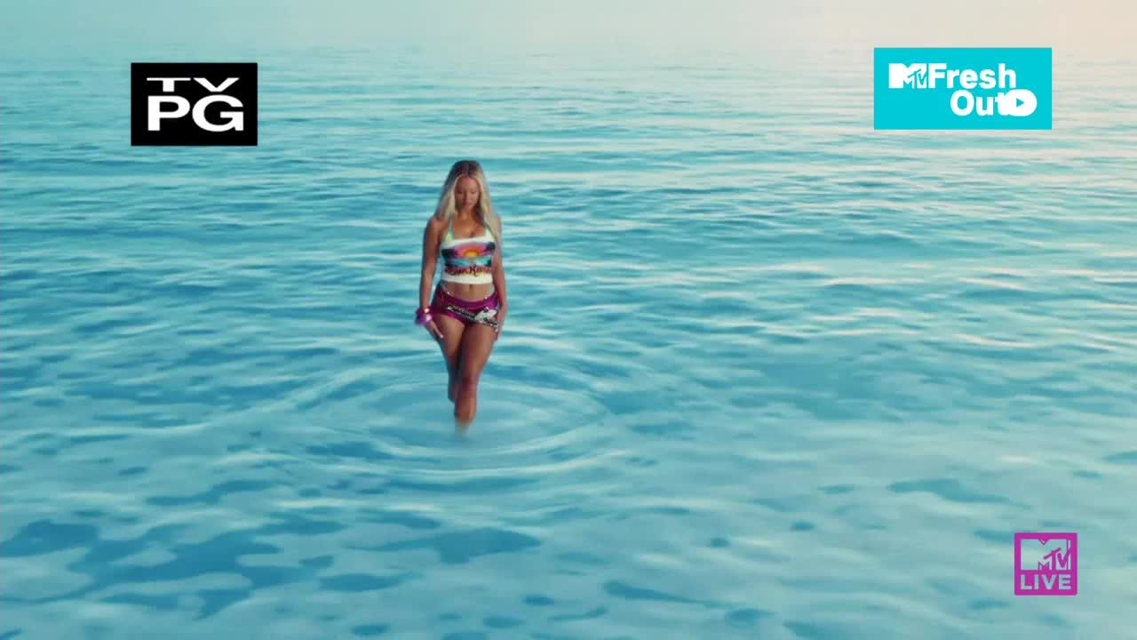 A woman in a bikini walks toward the viewer through shallow water, the waves barely reaching her knees. The scene is set on a bright day, and the MTV Live logo is visible in the corner.
