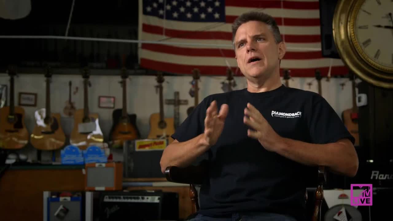 The man in the black t-shirt gestures with his hands as he speaks. Behind him, a wall of guitars and a large American flag hang in the background.
