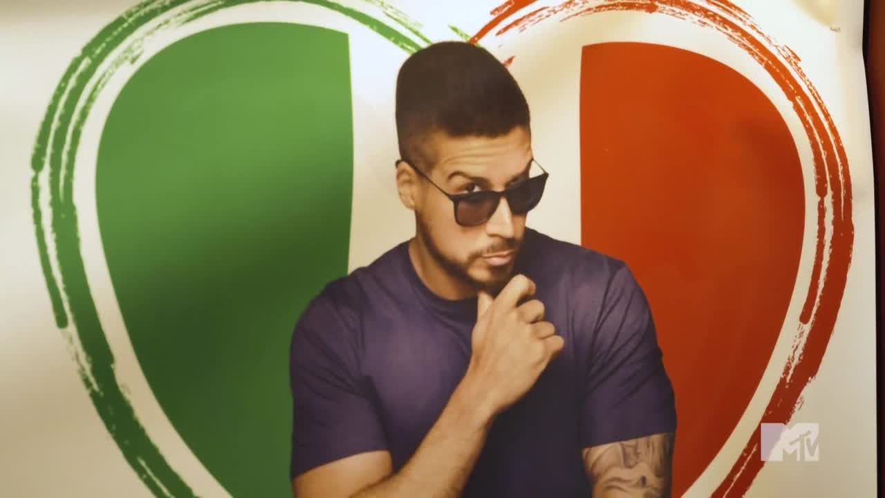 A man in sunglasses and a purple shirt poses in front of a heart-shaped Italian flag. He's holding his chin, looking directly at the camera with a slight smirk.
