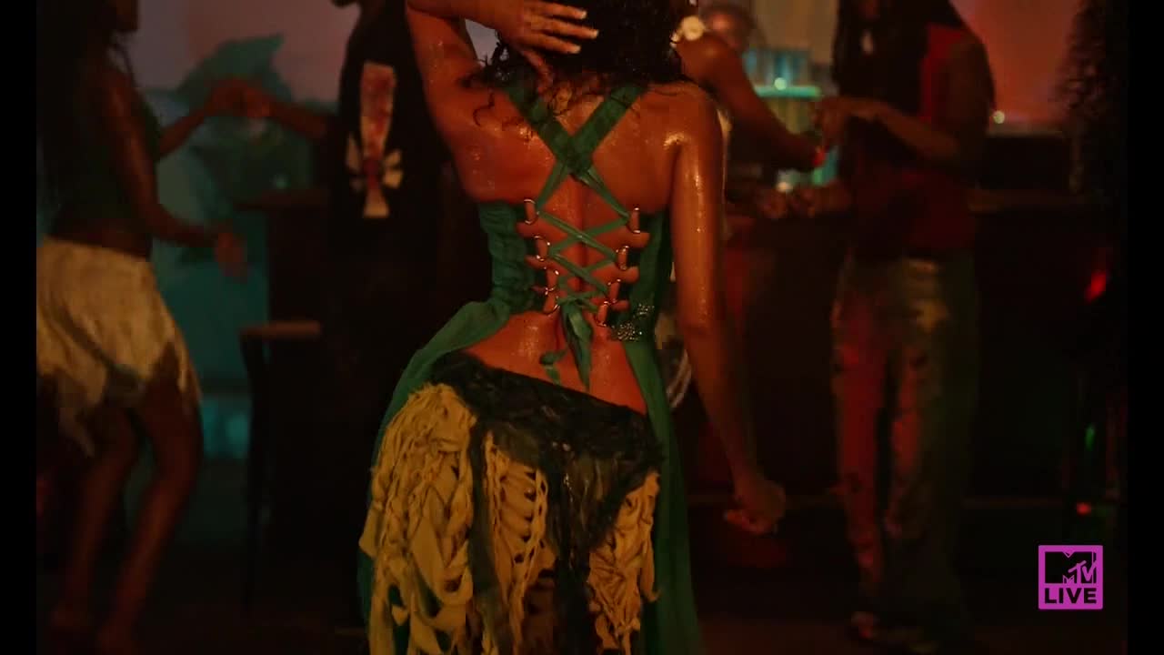 The dancer's back glistens as she moves, the green corset laced up tight. Other people sway nearby, the MTV Live logo visible in the corner.
