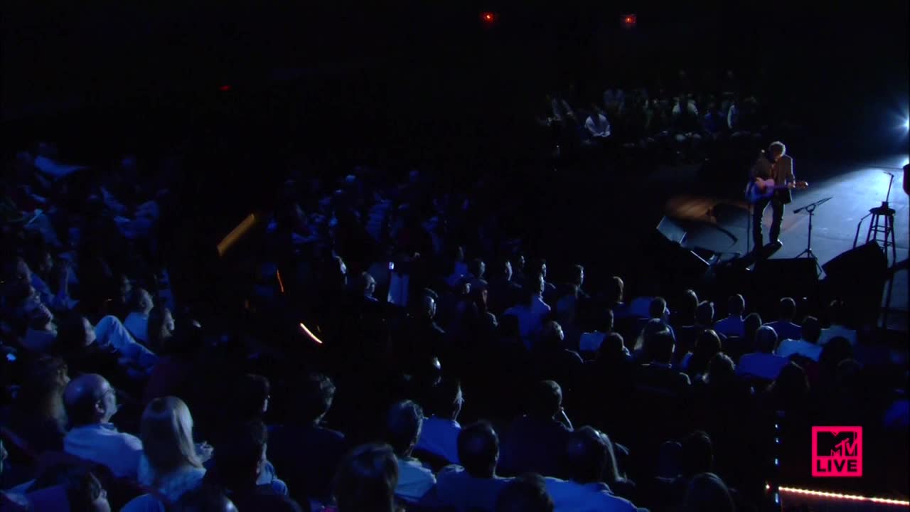 A musician stands alone on a brightly lit stage, strumming a guitar. The audience is mostly in shadows, but you can see their faces as they watch the performance on MTV Live.
