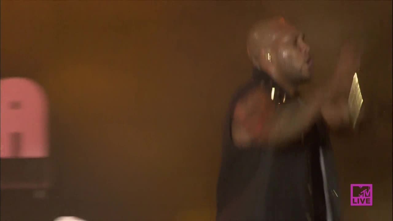 The performer is bathed in warm light, gesturing emphatically with both hands. His bald head glistens as he sings, and the MTV Live logo is visible in the corner.
