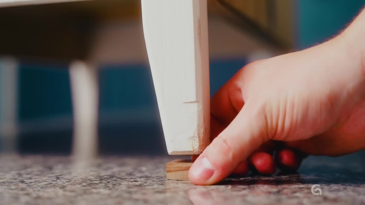 A hand carefully positions a small wooden block beneath a white painted table leg. The leg rests on the block, seemingly leveling the furniture.
