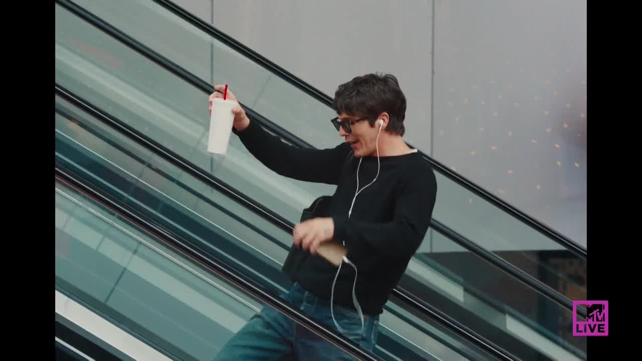 A man with headphones and sunglasses rides the escalator, holding a drink aloft as he seems to dance. He's got a backpack and a phone in his other hand.
