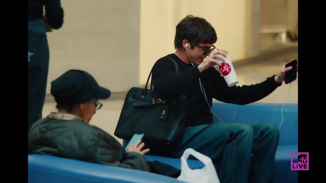 A man in a black sweater sips from a Wendy's cup while holding a phone, his wired earbuds dangling. Another person, wearing a cap and glasses, looks at their phone on the blue bench beside him.
