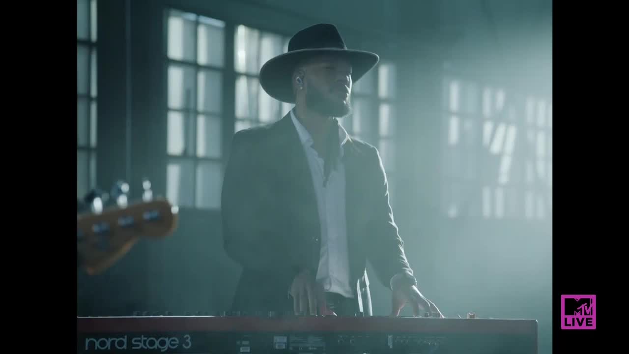 A man in a hat and suit is playing the keys on a red keyboard. The scene is dimly lit, with a bass guitar peeking in from the side.
