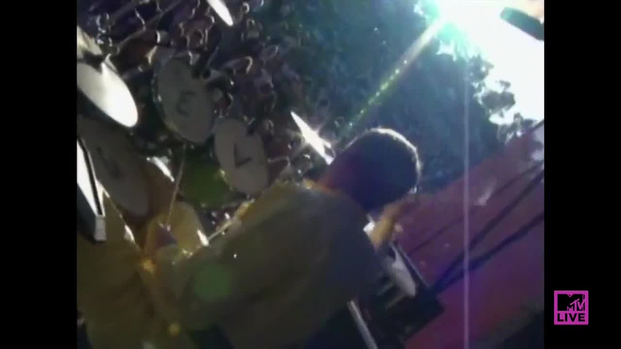 The drummer, bathed in sunlight, is hammering away at a kit surrounded by cymbals. Bright light flares from above as the sticks blur with each powerful strike.

