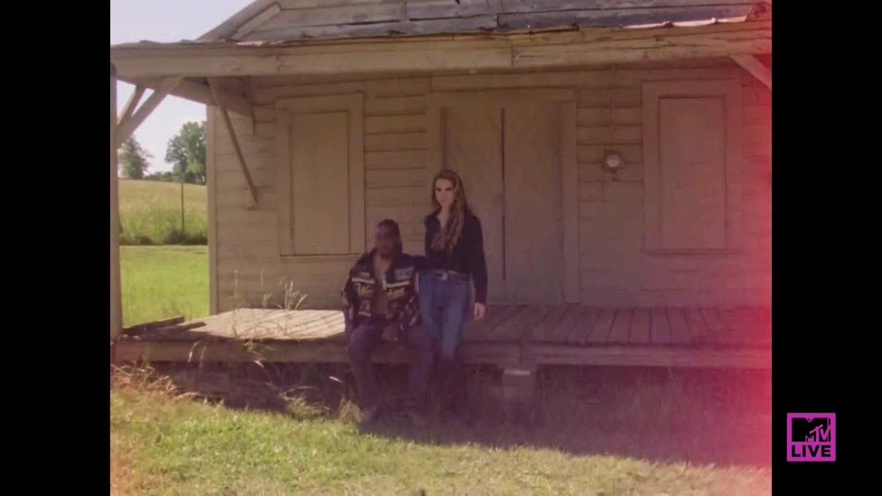 A man in a leather jacket sits on the porch of a weathered building, while a woman in a black jacket stands beside him. The MTV Live logo is visible in the corner, and the scene is bathed in a warm, hazy light.
