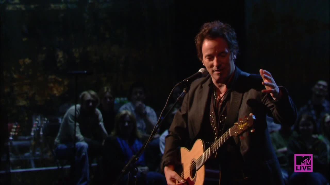 Bruce Springsteen strums an acoustic guitar, his voice amplified by a microphone as he sings. He gestures with his right hand, connecting with the audience seated in the background.
