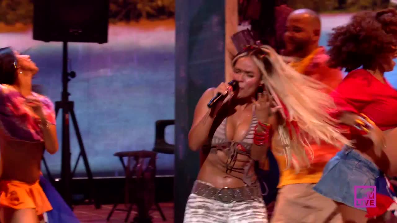 A woman in a halter top sings into a microphone, her long blonde hair flowing. Dancers in colorful outfits move energetically around her on a stage.
