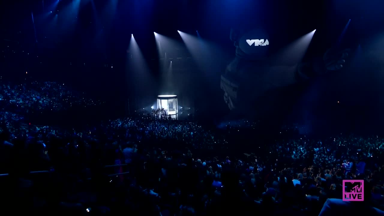 The crowd is packed, their phones held high, illuminated by the stage lights. A small, glowing structure sits center stage, with figures inside.
