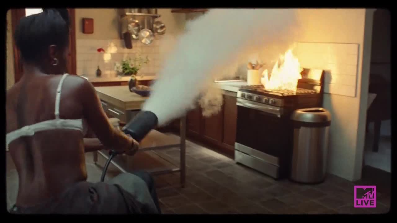 A woman in a bra aims a fire extinguisher at a stove engulfed in flames. Thick white smoke billows out, obscuring the kitchen.
