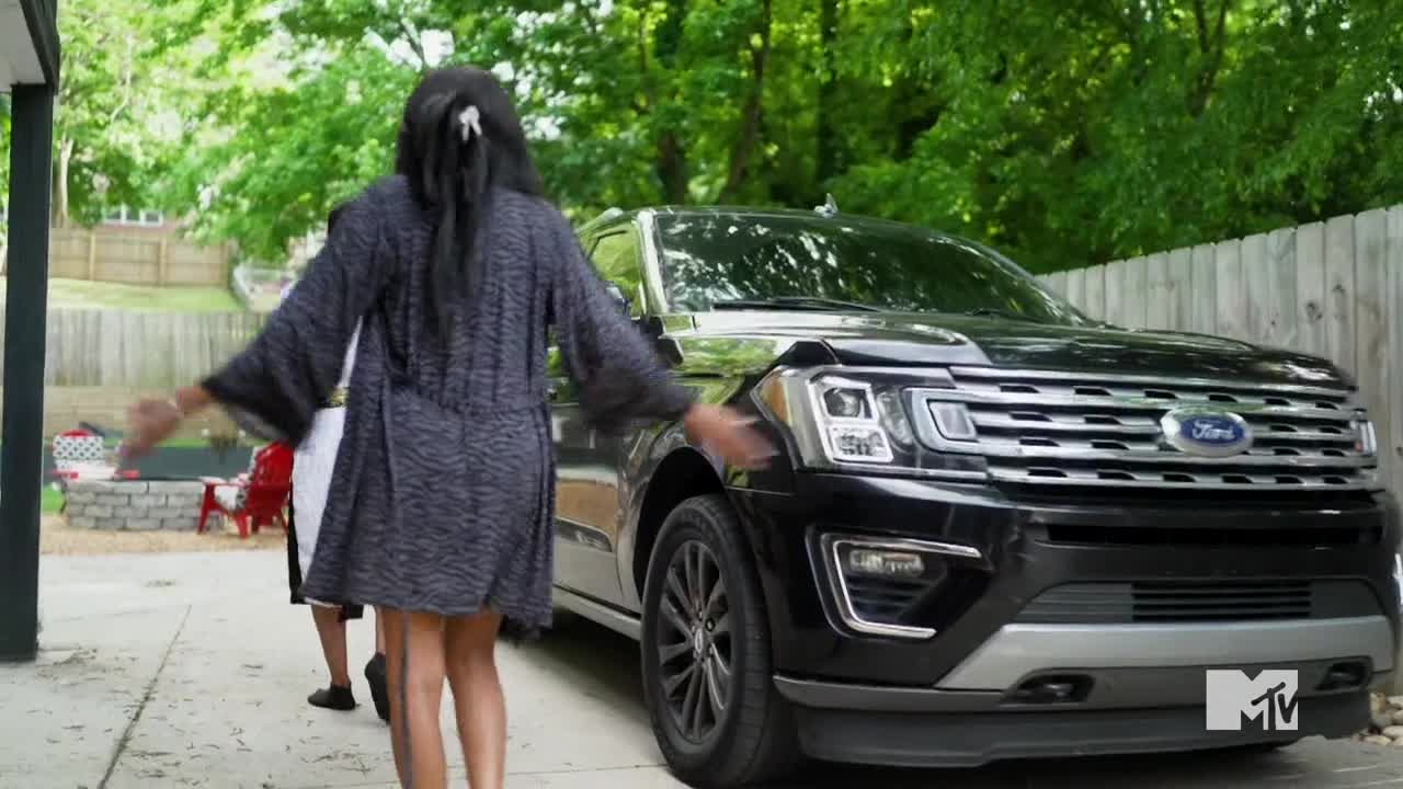 A woman in a patterned robe approaches a black SUV, her arms outstretched. The MTV Live logo is visible on the car's front bumper.
