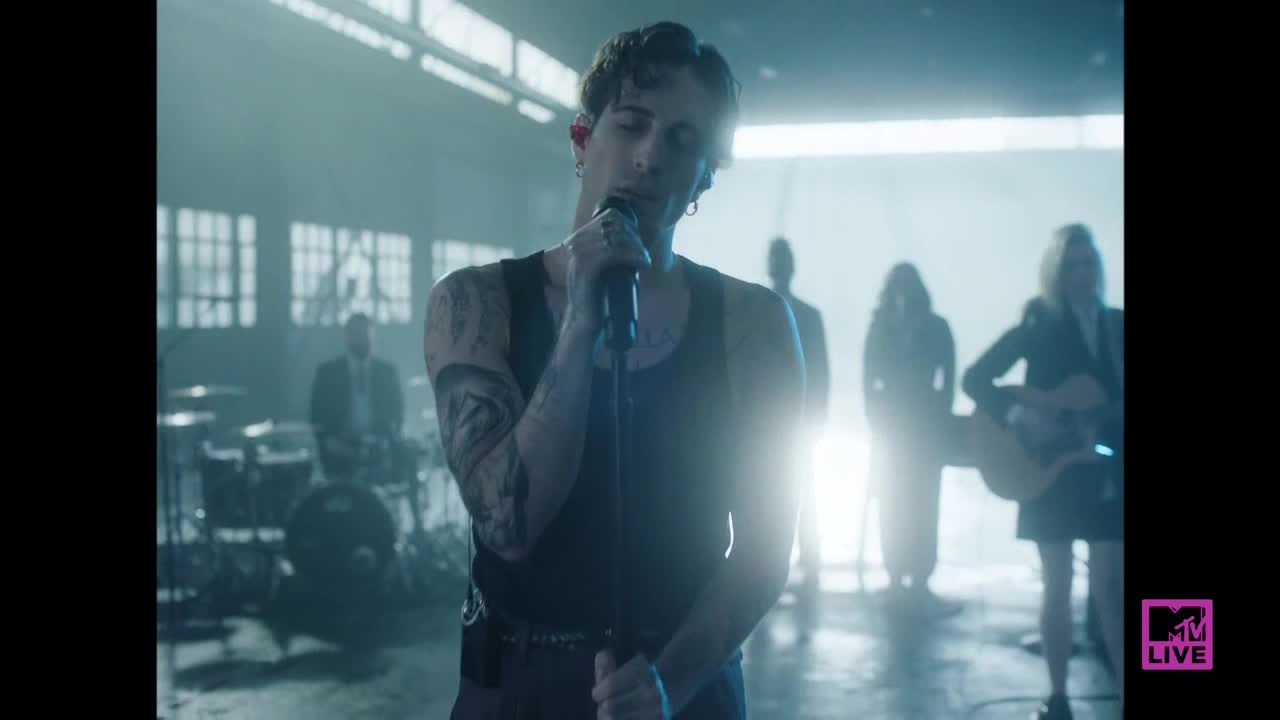 The singer, in a black tank top, is holding a microphone, his tattooed arm visible. Behind him, the band members are silhouetted in the dim, hazy light.
