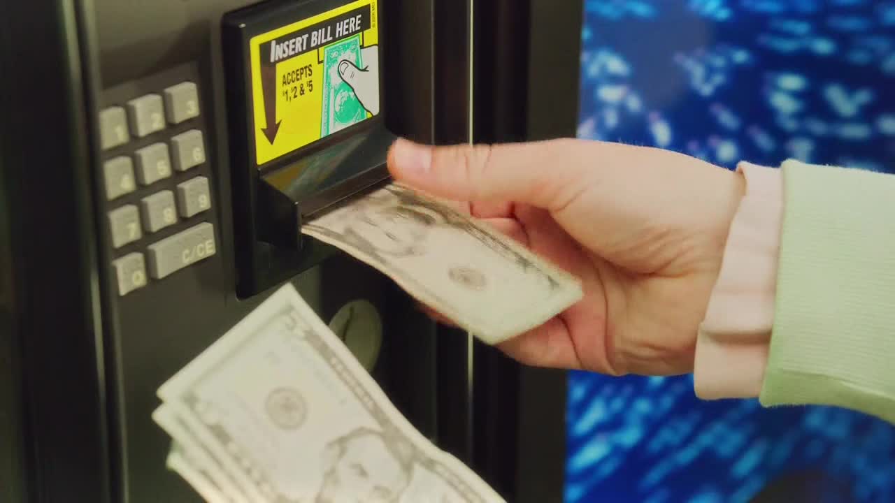 A hand slides a five-dollar bill into a vending machine's slot. More cash rests in the person's other hand, ready to go in next.
