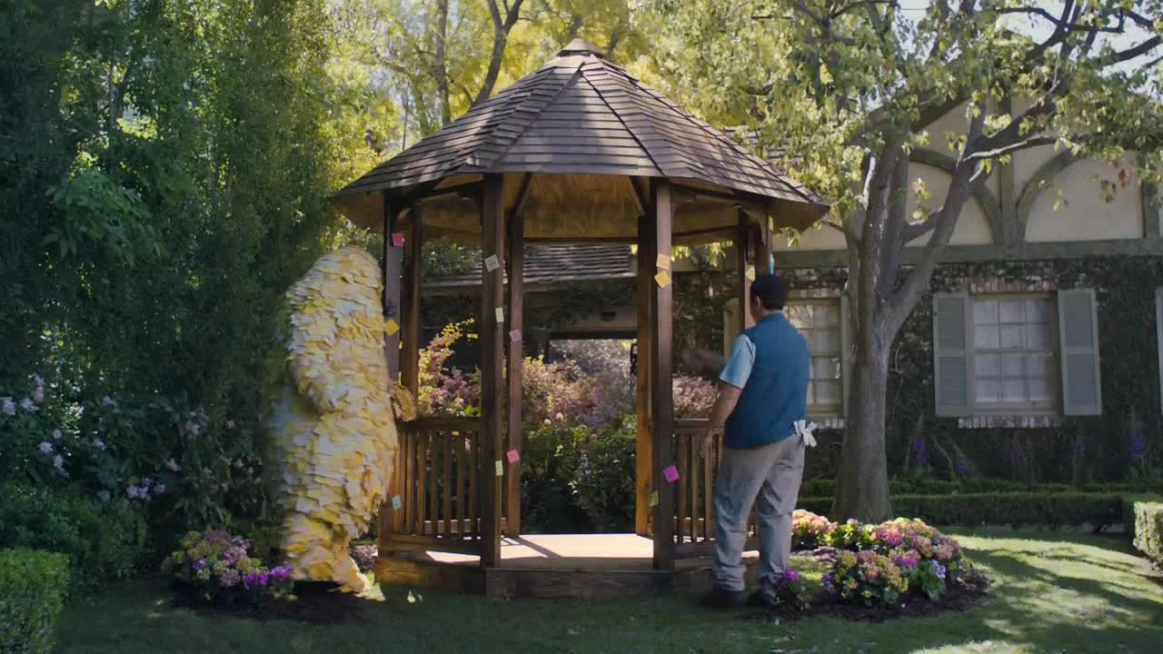 A person in a blue vest and gray pants stands inside a gazebo, looking out at a large yellow figure covered in what looks like sticky notes. The scene takes place in a well-manicured garden, with a house visible in the background.
