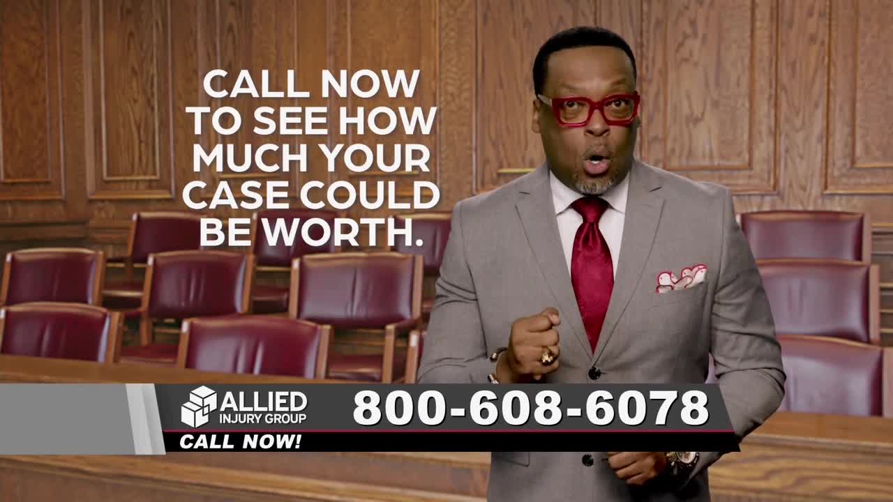 A man in a suit, with bright red glasses and tie, gestures emphatically toward the camera. He's standing in what looks like a courtroom, with a phone number displayed below.
