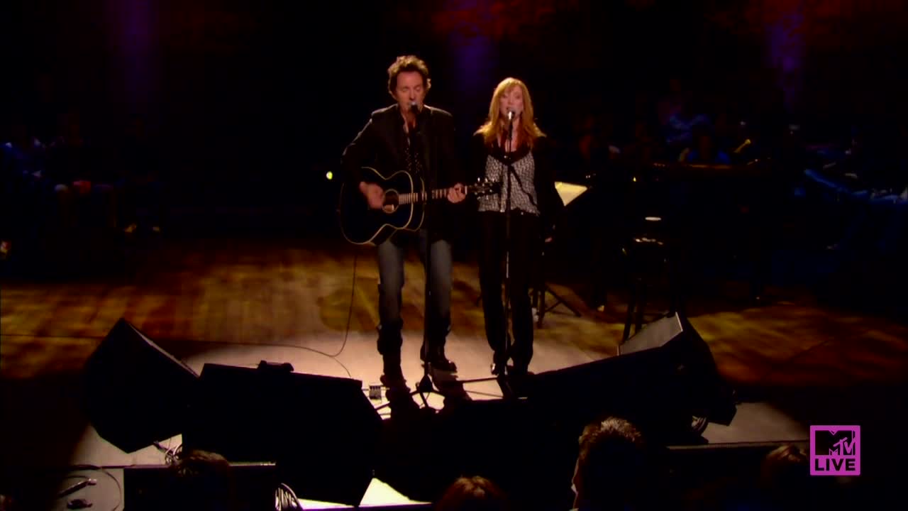 A man strums an acoustic guitar, singing into a microphone. Beside him, a woman with long red hair also sings, both illuminated by spotlights on a stage.
