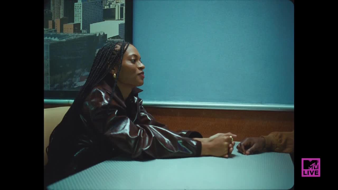A woman in a shiny, maroon jacket sits at a table, her hands clasped in front of her. Across from her, another person's hand rests on the table, the scene framed by a backdrop of a cityscape.
