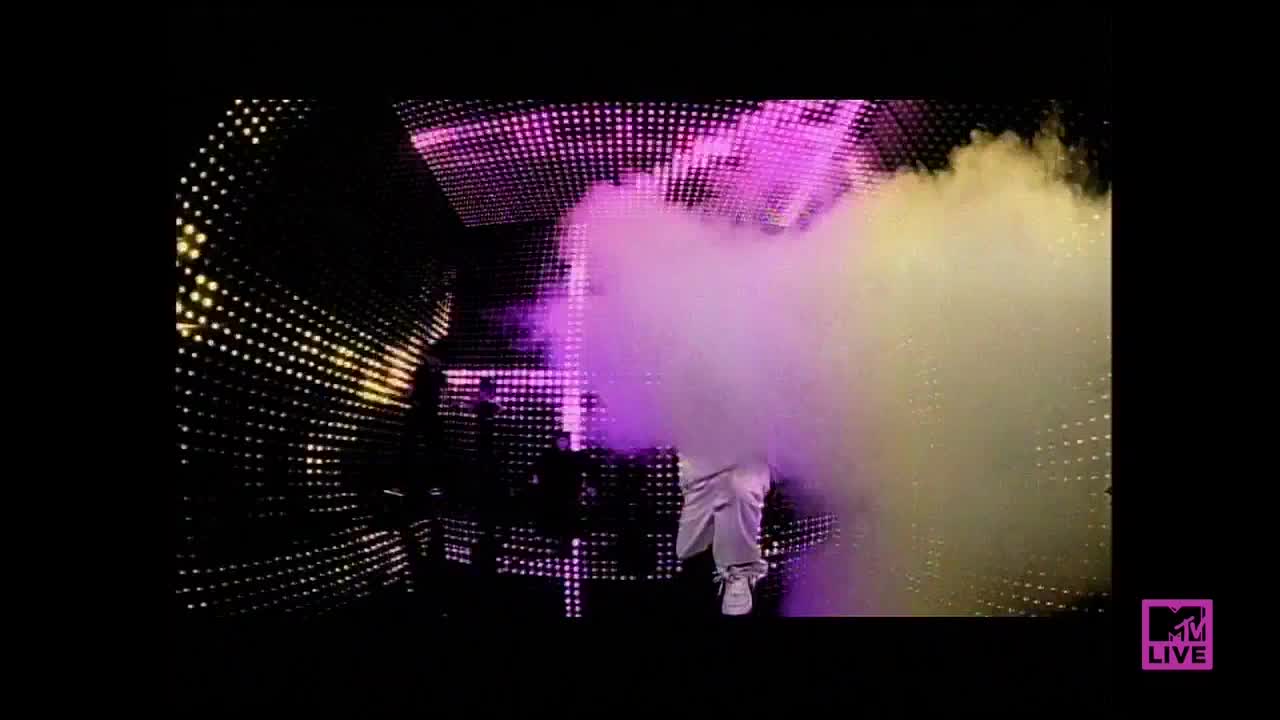 A figure in white pants and sneakers is engulfed in a cloud of smoke, illuminated by the stage lights. The backdrop is a wall of glowing dots, with the MTV Live logo visible in the corner.
