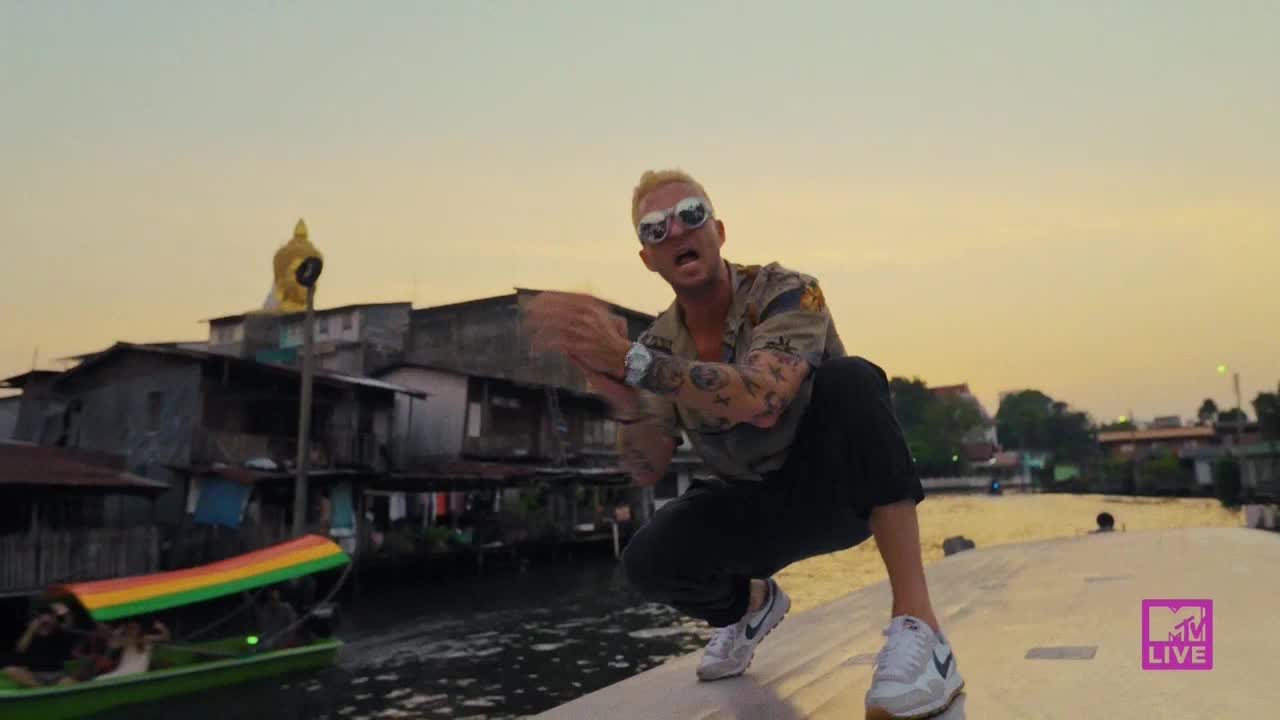 A man in sunglasses squats on a concrete ledge, clapping his hands. Behind him, a brightly colored boat glides along a canal, and the MTV Live logo is visible in the corner.
