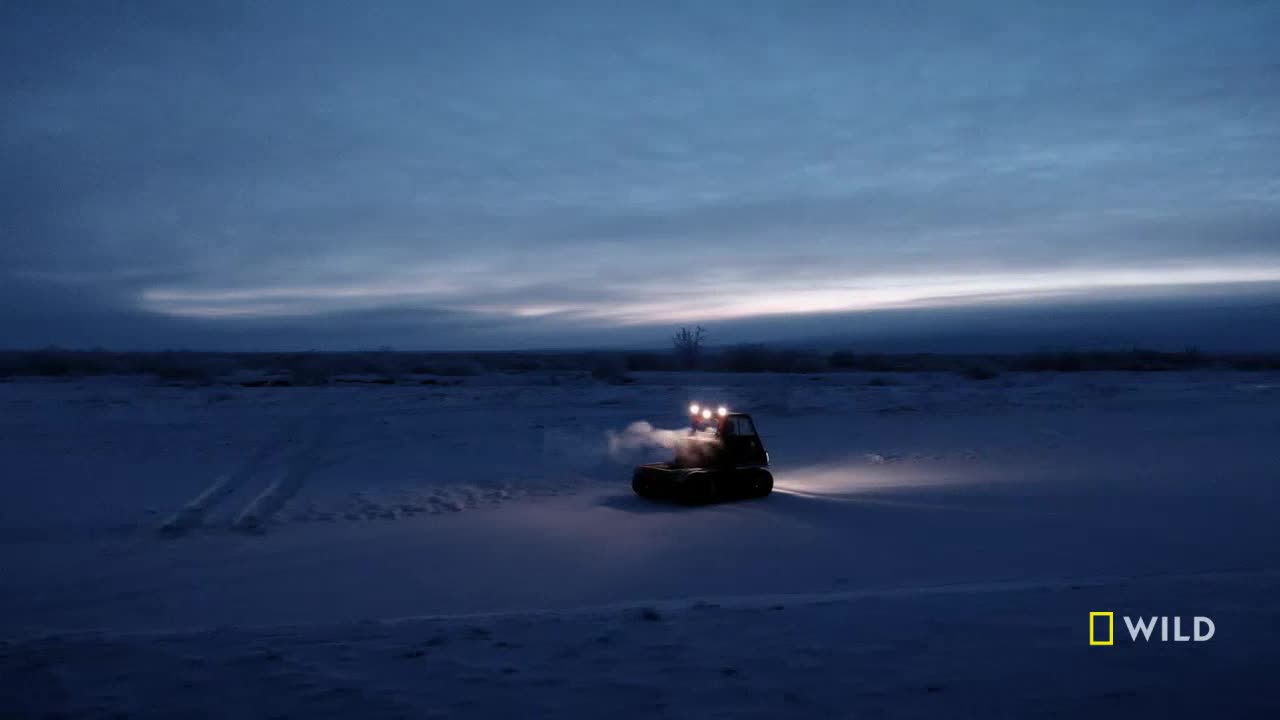 A small, tracked vehicle moves across a snowy expanse, its headlights cutting through the dim light. Steam rises from its engine as it heads towards the horizon.
