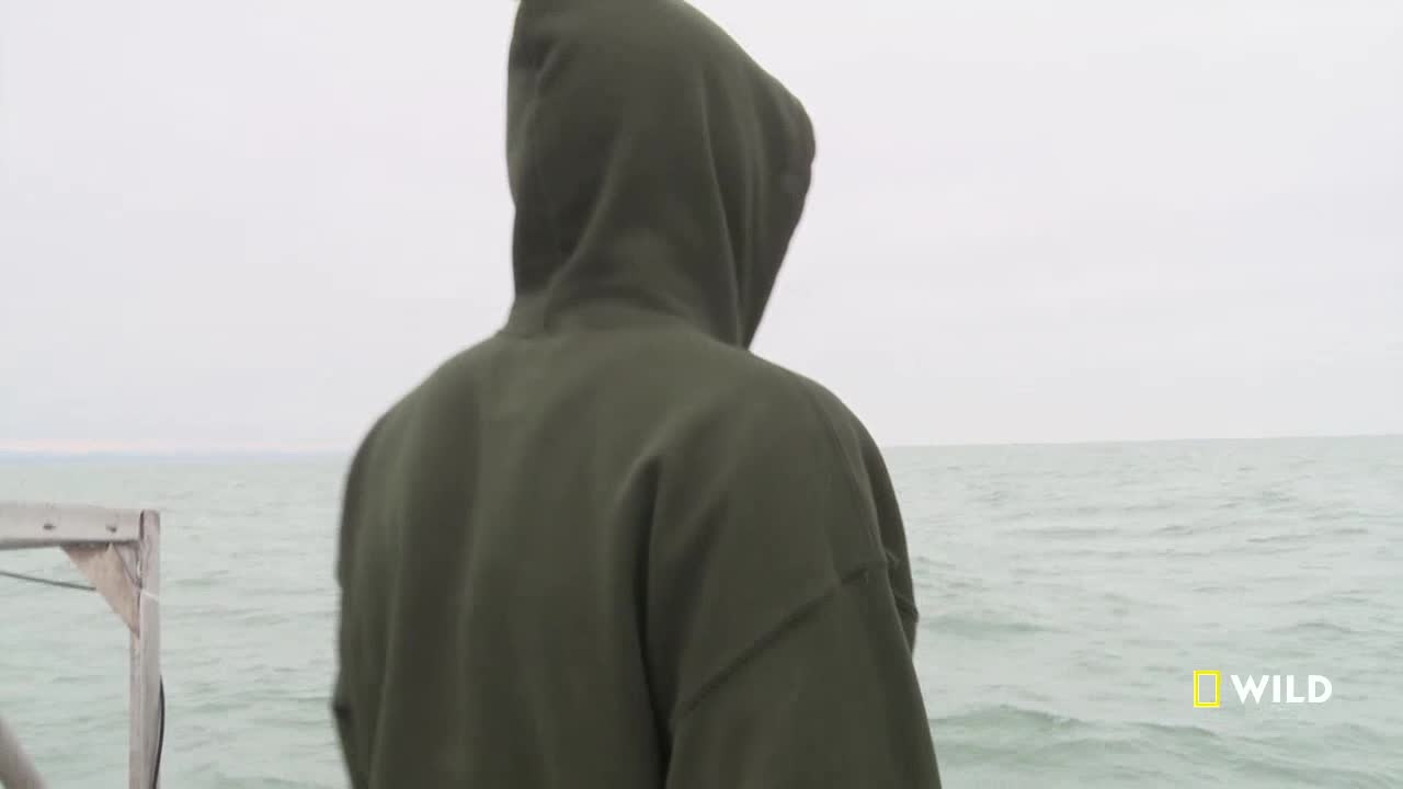 A person in a hooded sweatshirt stands on a wooden structure, gazing out at the choppy water. The sky is overcast, and the horizon blends into the sea.
