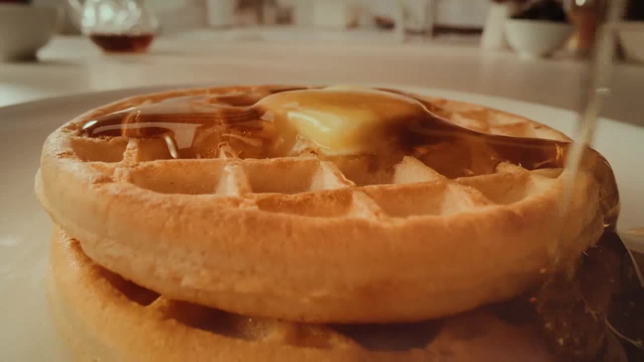 A pat of butter sits melting atop a stack of golden waffles. Amber syrup streams down, pooling in the waffle's squares.
