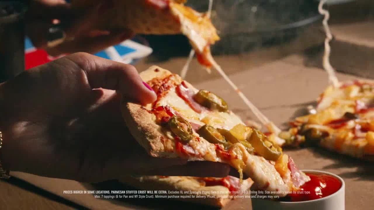 A hand with pink nail polish holds a slice of pizza, the cheese stretching into long, gooey strands.  Jalapeños and other toppings are visible on the pizza, and a small dish of red sauce sits nearby.
