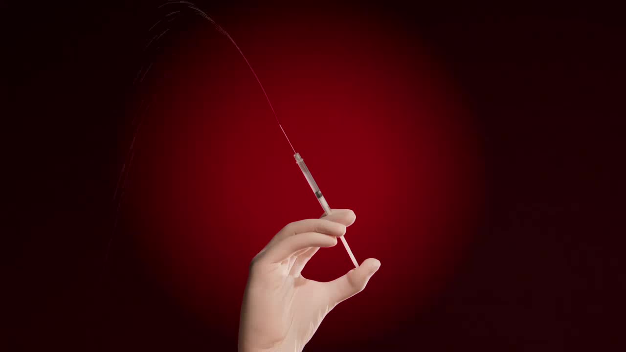 A hand holds a syringe, and a thin stream of red liquid arcs away from the needle.  The background is a deep, saturated red.
