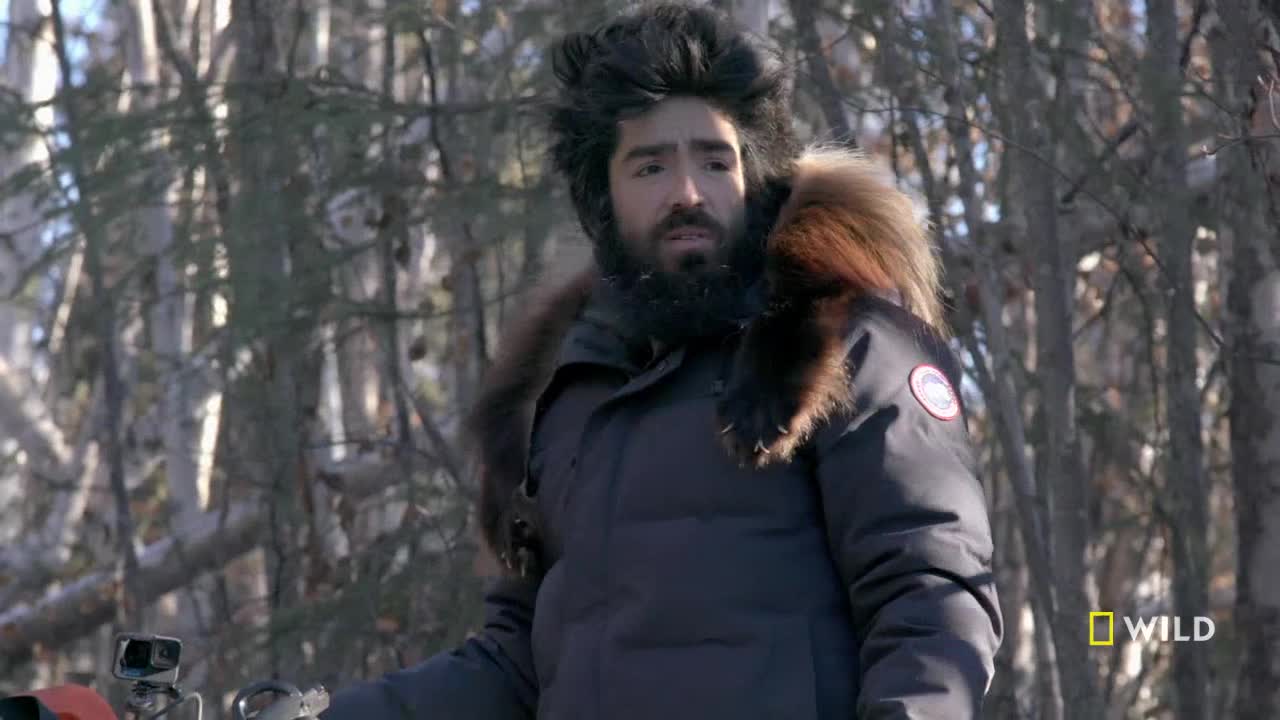 A man in a fur-trimmed parka stands in a snowy forest, looking slightly perplexed. He's on location for National Geographic Wild, likely filming a nature documentary in the United States.
