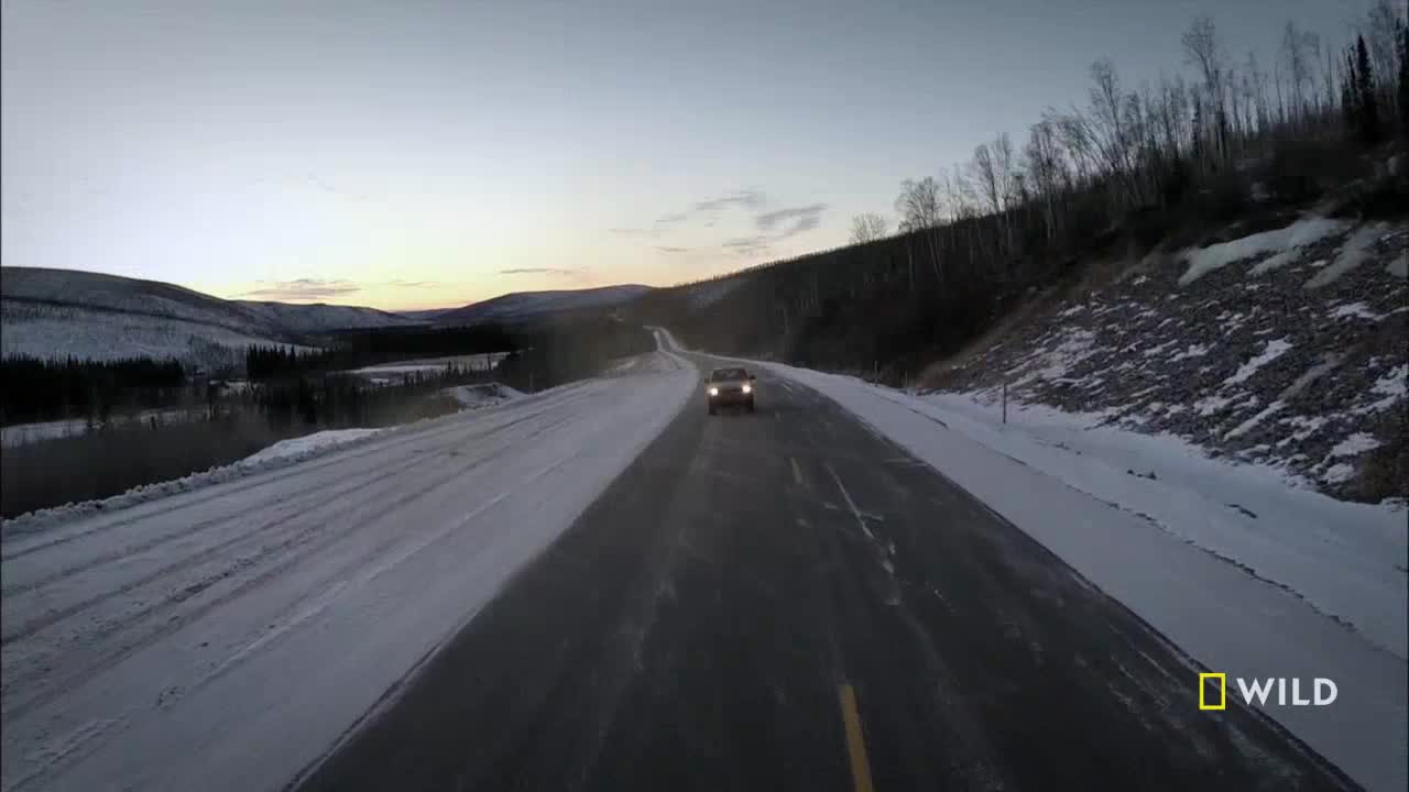 A vehicle, headlights illuminated, approaches on a snow-lined road in the United States. The sky is a soft, dusky blue as the car travels down the long, winding highway.
