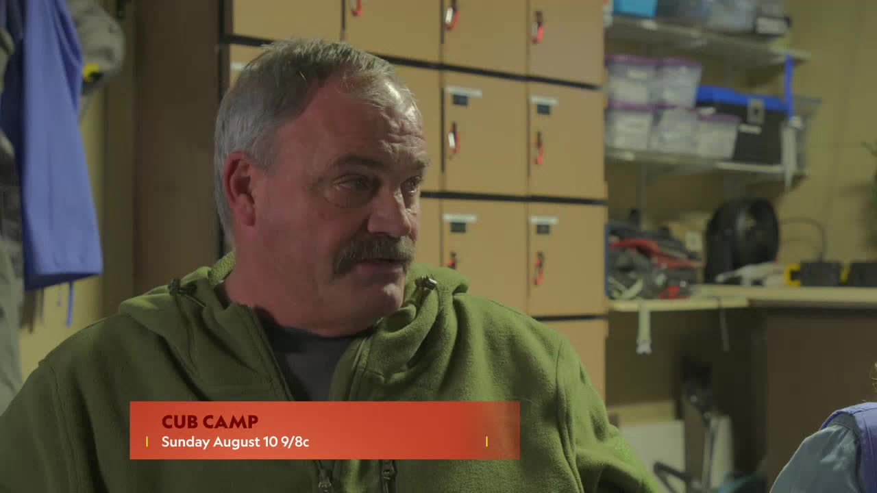A man with a thick mustache is speaking, his gaze directed off-camera.  Behind him, a stack of boxes and a shelf filled with supplies are visible.
