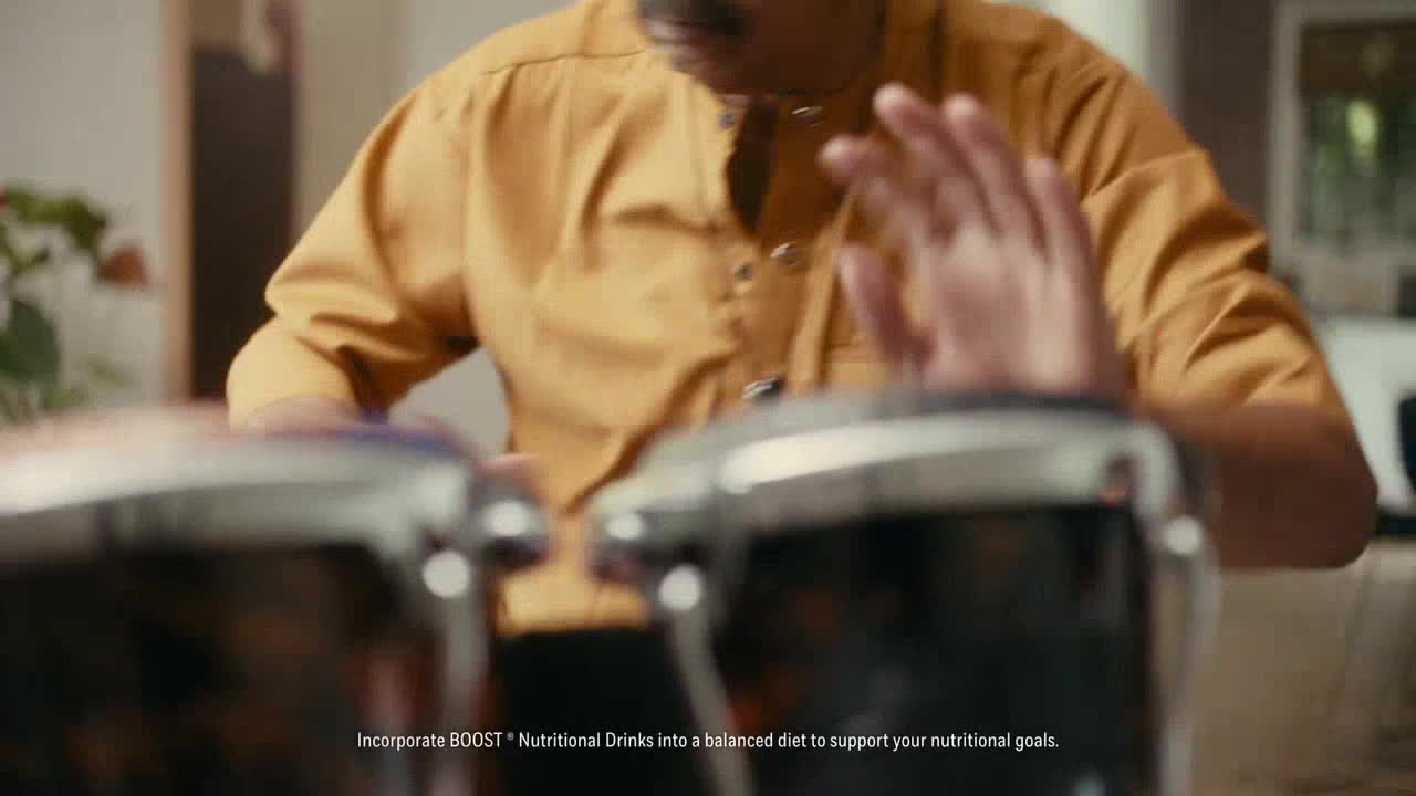 A man in a yellow shirt is drumming on a pair of shiny bongos. His hands are raised, about to strike the drums.
