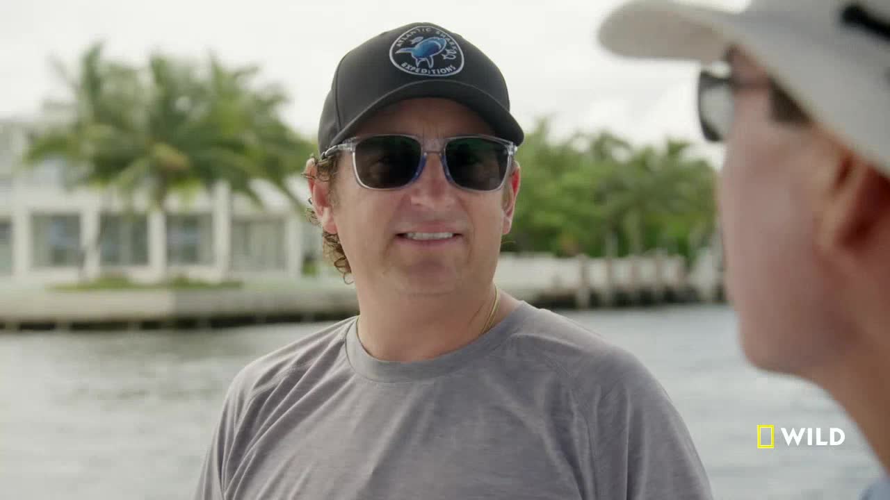 A man in a cap and sunglasses is talking to someone out of frame, a waterfront in the background. The National Geographic Wild logo is visible in the bottom right corner.
