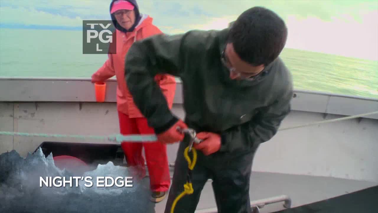 On a boat, a man in a green jacket and red gloves is working with a metal clip and a rope. A person in an orange jacket and red pants stands nearby, holding a cup.
