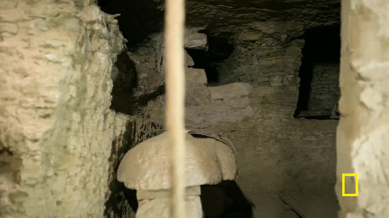 A thick, pale rope hangs down, obscuring the view of a rough-hewn stone chamber. The walls are made of stacked blocks, and a dark opening suggests a deeper passage.
