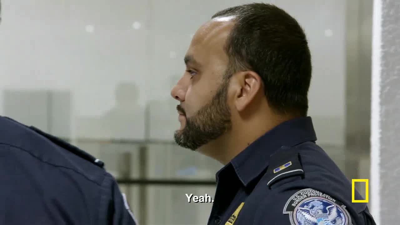 A U.S. Customs and Border Protection officer, seen from the side, looks toward something outside of the frame. The word "Yeah" appears on the screen, likely a snippet from a National Geographic documentary.
