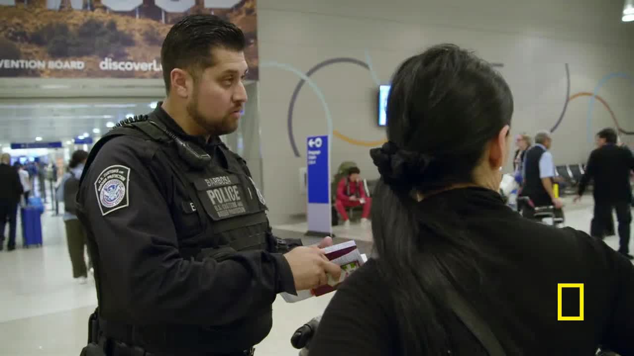 A U.S. Customs and Border Protection officer, in uniform, holds a passport and looks at a traveler. The woman in front of him has a National Geographic logo on her back.
