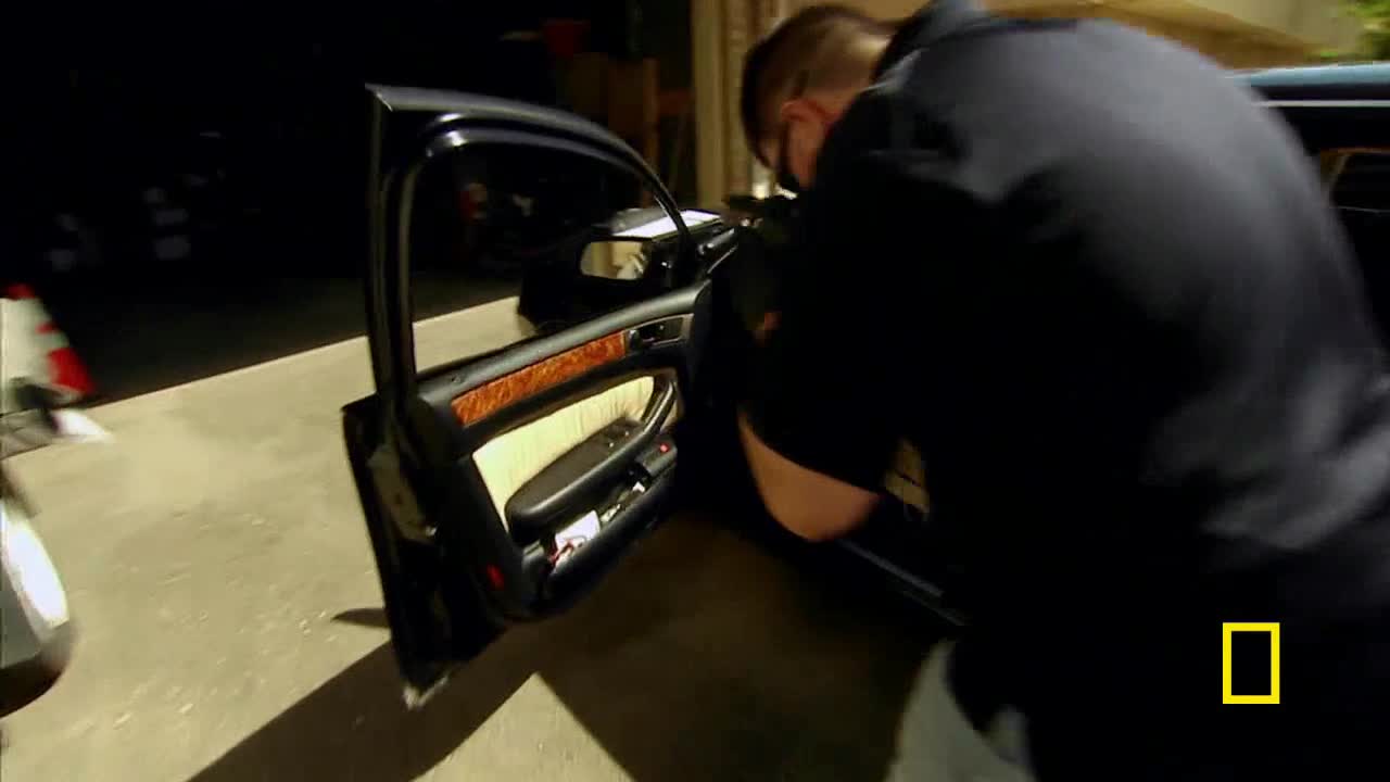 A man in a dark shirt is leaning into the open door of a black car, his back to the camera. The car's interior has wood trim and light-colored seats.
