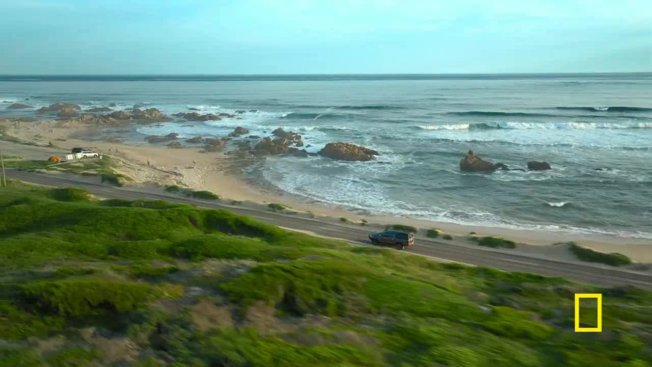 A dark SUV drives along a coastal road, the ocean waves crashing against the rocky shore. The scene is framed by green grassy hills and a clear, hazy sky.
