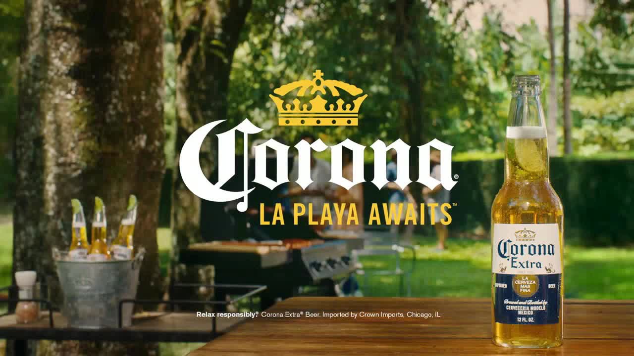 A bottle of Corona Extra sits on a wooden table, its label clearly visible. In the background, people gather around a grill, enjoying a sunny outdoor setting.
